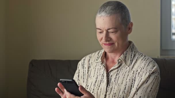 Uma mulher idosa olha para a tela do smartphone e sorri sinceramente. — Vídeo de Stock