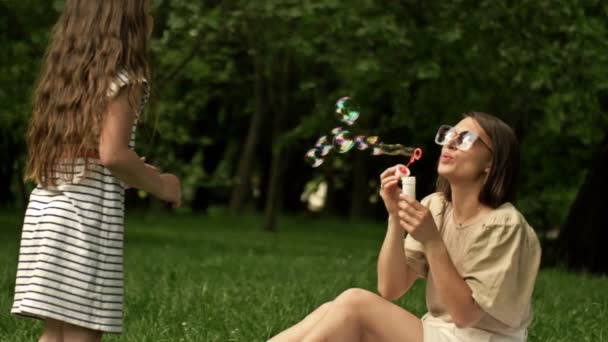 Beautiful young woman sits on green grass in the park and blows soap bubbles. Her 6-year-old daughter happily catches rainbow bubbles. Family holiday. — Stock Video