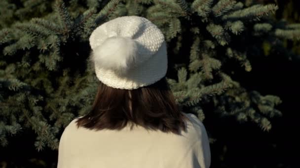 Chica en un sombrero blanco de punto admira un abeto verde esponjoso. Vista trasera. — Vídeo de stock