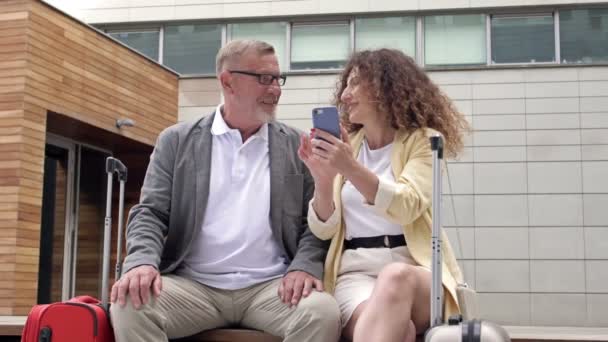 Krásný dospělý pár s kufry sedí na lavičce a čeká na svůj let. Muž a žena byli pobaveni tím, co viděli na obrazovce mobilního telefonu. — Stock video