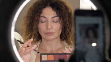 İyi bakımlı, orta yaşlı güzellik blogcusu göz makyajından bahsediyor. Aynanın önünde oturan bir kadın ve bir video kamera..