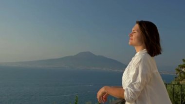Genç bir kadın güzel bir deniz ve dağ manzaralı bir balkonda duruyor. Kadın mutlu bir şekilde yüzünü güneş ışınlarına maruz bırakıyor..