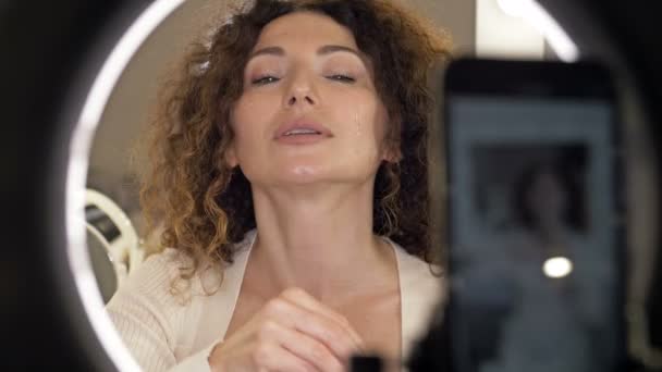 Pozytywna, piękna kobieta w średnim wieku demonstruje zastosowanie nawilżającego i odżywczego produktu kosmetycznego do pielęgnacji skóry. Beauty blogger obecny kosmetyki uroda siedzi przed kamerą dla — Wideo stockowe