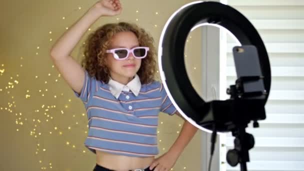 Концепция молодого блогера. Веселая девочка-подросток танцует перед камерой, снимая видео с помощью телефона на штативе дома, создавая свой модный контент в мобильном приложении, чтобы поделиться им в социальных сетях. — стоковое видео