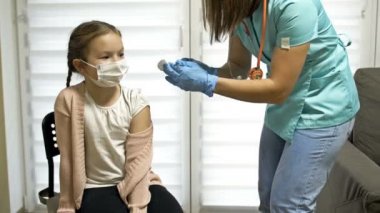Koruyucu üniformalı ve tıbbi maskeli kadın hemşire klinik laboratuvar odasında 7-8 yaşındaki kıza kovid 19 aşısı yapıyor..