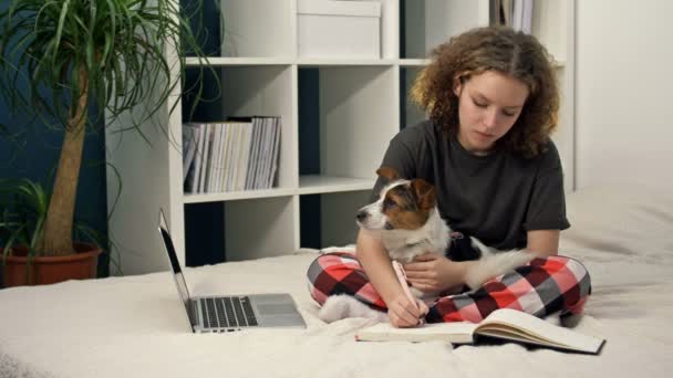 Afstandsonderwijs tijdens COVID-19. Tienermeisje die huiswerk maakt zittend op het bed met een computer. Ze houdt haar geliefde hond in haar armen.. — Stockvideo