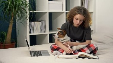 COVID-19 sırasında uzaktan öğrenme. Ödev yapan genç bir kız bilgisayarla yatakta oturuyor. Sevgili köpeğini kollarında tutuyor..
