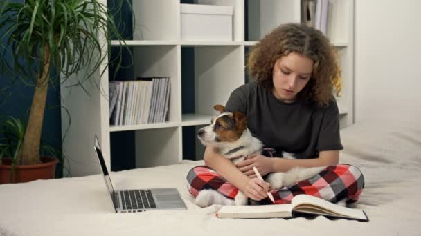 Afstandsonderwijs tijdens COVID-19. Tienermeisje die huiswerk maakt zittend op het bed met een computer. Ze houdt haar geliefde hond in haar armen.. — Stockvideo