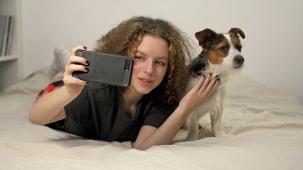 Beautifu teenage girl ltake a photo selfie bu mobile phone with her cute dog in cozy bedroom. — Stockvideo