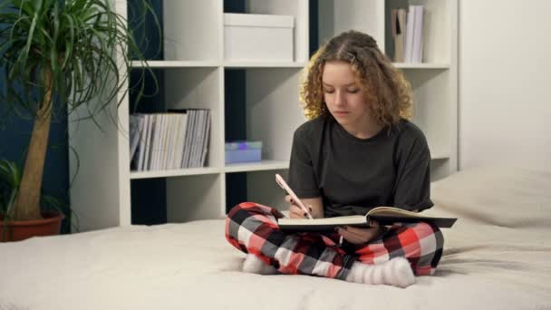 Słodka nastolatka odrabia lekcje siedząc w domu na łóżku. Młoda piękna dziewczyna myśli i pisze coś w notatniku, studiuje online, uczenie się na odległość, samokształcenie. — Wideo stockowe