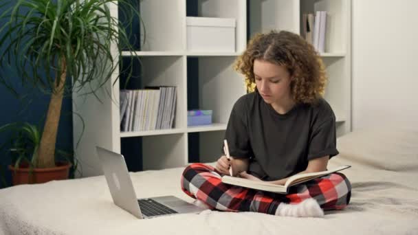 귀여운 십 대 소녀 가집 침대에 앉아서 숙제를 하고 있다. 한 젊고 아름다운 소녀가 생각하고 노트에 뭔가를쓰고, 온라인으로 공부하고, 거리를 배우고, 스스로 배우고,. — 비디오
