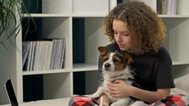 COVID-19 sırasında uzaktan öğrenme. Ödev yapan genç bir kız bilgisayarla yatakta oturuyor. Sevgili köpeğini kollarına alır, ona sarılır ve onu öper..