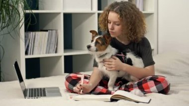 COVID-19 sırasında uzaktan öğrenme. Ödev yapan genç bir kız bilgisayarla yatakta oturuyor. Sevgili köpeğini kollarında tutuyor..