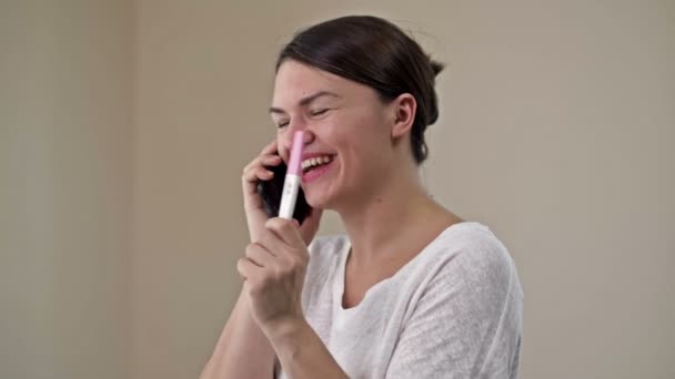 Happy woman holding her positive result on pregnancy test and talking by a smartphone with the family. — Stock Video