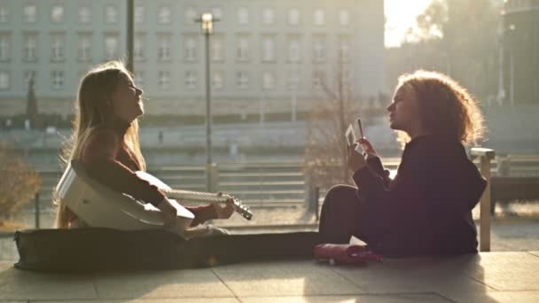 Adolescentes sentadas en el terraplén. Uno toca la guitarra, el otro arregla su maquillaje. Ocio adolescente. — Vídeos de Stock