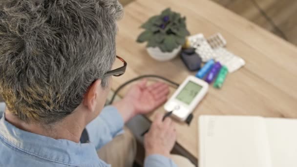 En äldre kvinna mäter sitt blodtryck och skriver ner resultaten i en anteckningsbok. Övervakning i hemmet. — Stockvideo