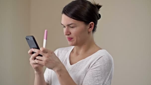 Ung kvinna är glad att se ett positivt graviditetstest. Hon skyndar sig att dela sin glädje på telefonen med en nära och kära. — Stockvideo