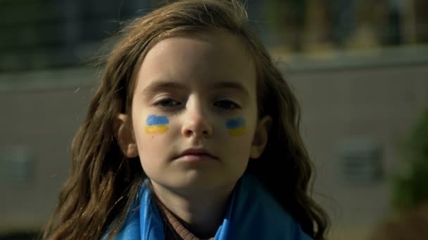 En liten flickas ansikte. Barnen kinderna är målade med de gula och blå färgerna i den ukrainska flaggan. Barn ber om fred. — Stockvideo
