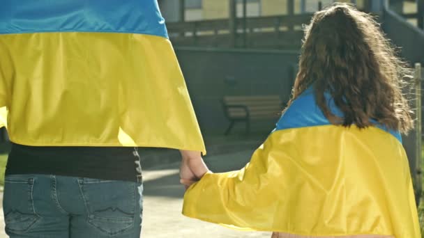 Дорослий чоловік тримає руку маленької дівчинки. Плечі обох сторін покриті прапором України. Погляд назад. — стокове відео