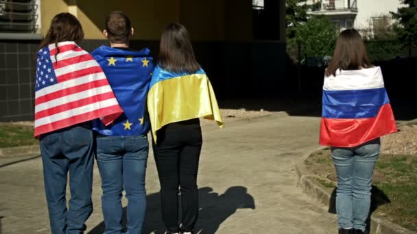 Groep omhelzende mensen onder de vlag van Oekraïne, de VS en de EU. Een symbool van solidariteit en steun. Een vrouw met een Russische vlag staat alleen. — Stockvideo