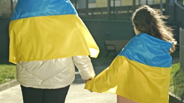 Een volwassen vrouw houdt de hand van een klein meisje vast. De schouders van beide zijn bedekt met de vlag van Oekraïne. Achteraanzicht. — Stockvideo