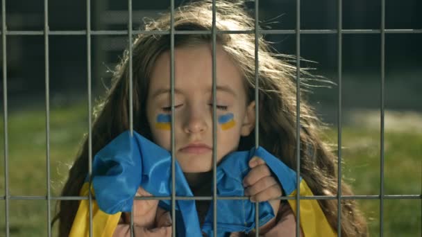 Klein meisje is bedekt met een Oekraïense vlag met haar ogen dicht achter een metalen rooster. Symbool van gebrek aan vrijheid. Oorlog in Oekraïne. — Stockvideo