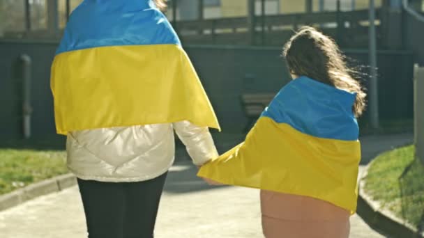 En vuxen kvinna håller en liten flickas hand. Axlarna på båda är täckta med Ukrainas flagga. Bakifrån. — Stockvideo