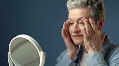 Yaşlı, kır saçlı bir kadın aynadaki yansımasını dikkatlice inceler. Yüzündeki kırışıklıklara hafifçe dokunur..