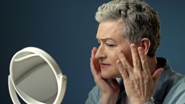 Una anciana mujer gris examina cuidadosamente su reflejo en el espejo, tocando ligeramente las arrugas en la piel de su cara. — Vídeo de stock
