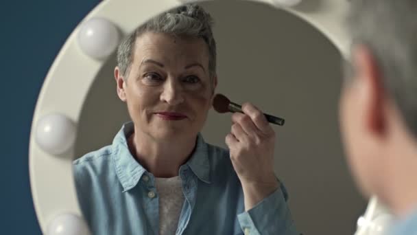 Mujer mayor bien arreglada aplica rubor en la mejilla con un cepillo de maquillaje, sentado solo en casa delante de un espejo. — Vídeos de Stock