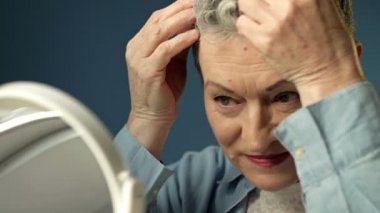 Gri saçlı yaşlı kadın evden çıkmadan önce saçlarını aynanın önünde düzleştiriyor..