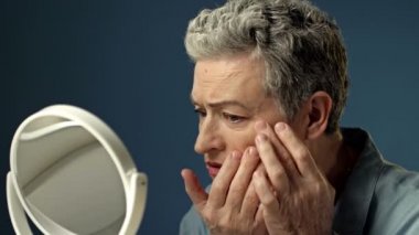 Yaşlı, kır saçlı bir kadın aynadaki yansımasını dikkatlice inceler. Yüzündeki kırışıklıklara hafifçe dokunur..
