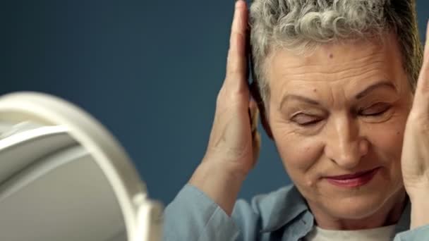 Γκρίζα ηλικιωμένη γυναίκα ισιώνει τα μαλλιά της μπροστά από έναν καθρέφτη πριν φύγει από το σπίτι.. — Αρχείο Βίντεο