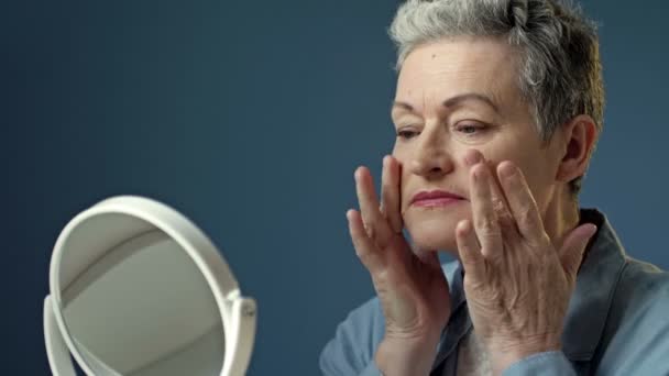 Eine ältere, ergraute Frau untersucht sorgfältig ihr Spiegelbild im Spiegel und berührt leicht die Falten auf der Haut ihres Gesichts.. — Stockvideo