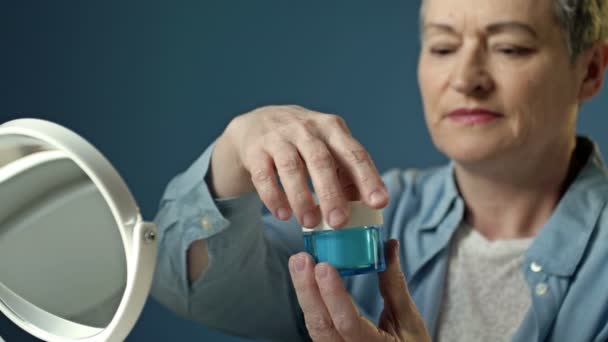 Una donna anziana applica una crema idratante anti-invecchiamento. Il pensionato bada alla sua apparizione, fa procedure cosmetiche. Utilizza la crema per il ringiovanimento, la rigenerazione, il sollevamento della pelle. — Video Stock