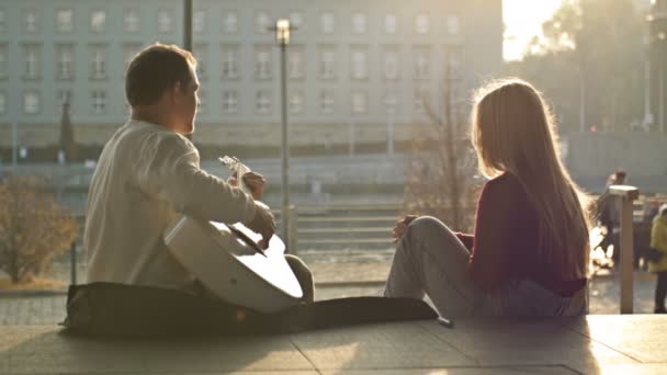 Data romântica à beira-mar. Alguns amantes estão sentados no dique da cidade. O tipo toca guitarra para a rapariga.. — Vídeo de Stock