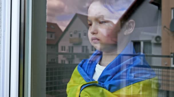 Kleines Mädchen mit der Flagge der Ukraine auf den Schultern blickt mit ernstem Gesichtsausdruck aus dem Fenster. — Stockvideo