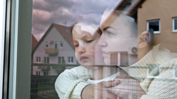 Mother and little daughter stand embracing and look sadly out the window. — Vídeos de Stock