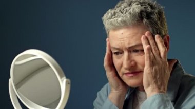 Yaşlı, gri saçlı bir kadın aynadaki yansımasını dikkatlice inceler. Yüzündeki kırışıklıklara hafifçe dokunur. Kadın üzgün..