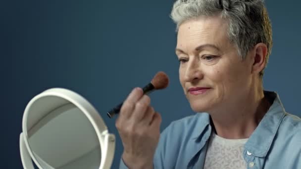 Ухоженная пожилая женщина наносит косметический продукт на лицо кисточкой для макияжа, сидя дома одна перед зеркалом. — стоковое видео
