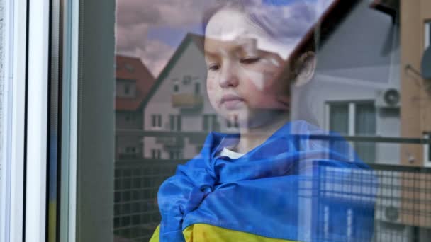 Kleines Mädchen mit der Flagge der Ukraine auf den Schultern blickt mit ernstem Gesichtsausdruck aus dem Fenster. — Stockvideo