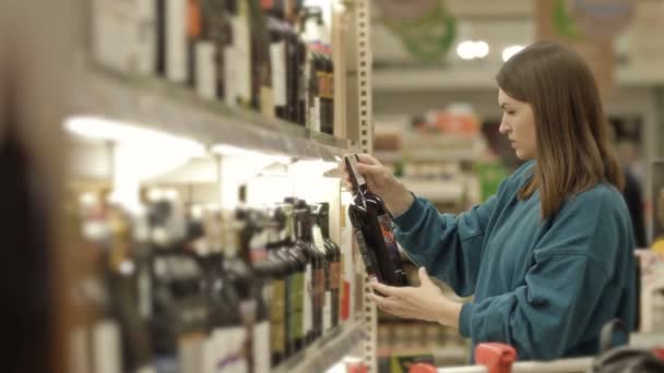 Genç bir kadın markette ya da içki dükkanında elinde alkolle rafların önünde beklerken şarabı seçiyor.. — Stok video
