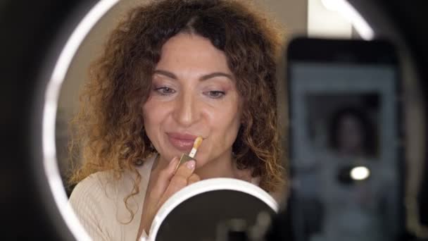 Schöner Schönheitsblogger mittleren Alters spricht über Lipgloss und legt ihn auf seine Lippen. Eine Frau sitzt vor einem Spiegel, der eine Videokamera reflektiert. — Stockvideo