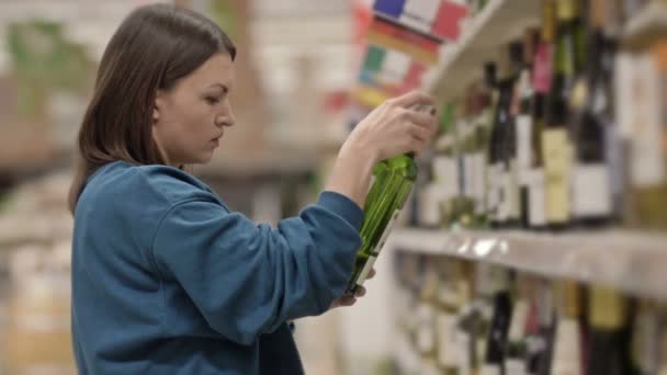 Mujer joven elige el vino de pie delante de los estantes con alcohol en un supermercado o licorería. — Vídeos de Stock