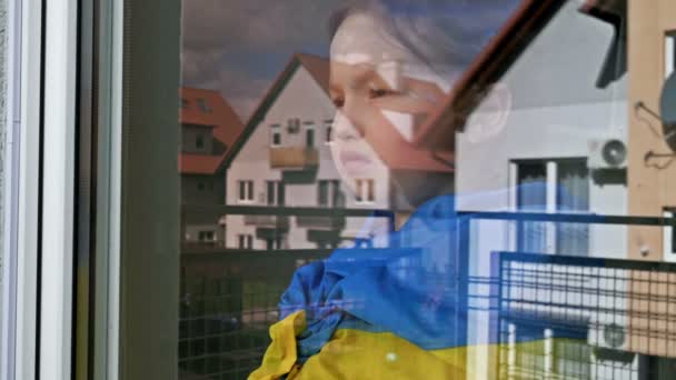 Κοριτσάκι με τη σημαία της Ουκρανίας στους ώμους της κοιτάζει έξω από το παράθυρο με μια σοβαρή έκφραση. — Αρχείο Βίντεο