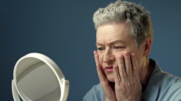 An elderly gray-haired woman carefully examines her reflection in the mirror, lightly touching the wrinkles on the skin of her face. Woman is upset. — Vídeos de Stock