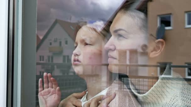 Mother and little daughter stand embracing and look sadly out the window. — стоковое видео