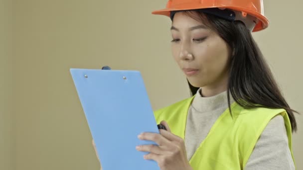 Asiático construtor mulher, vestindo roupas protetoras e um capacete, está escrevendo algo. — Vídeo de Stock