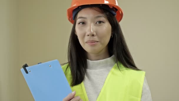 Asian woman builder in protective clothing. A young woman is holding a folder with documents and smiling. — Stockvideo
