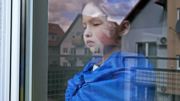 Niña con la bandera de Ucrania en los hombros mira por la ventana con una expresión seria. — Vídeos de Stock